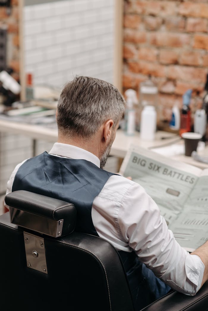 A senior man sits in a barbershop chair reading a newspaper, showcasing a relaxed moment.